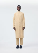 A person faces forward against a white background, wearing HOMME PLISSÉ ISSEY MIYAKE's TAILORED PLEATS 2 set in beige—featuring a long tailored jacket and matching pleated trousers—paired with black slip-on shoes.