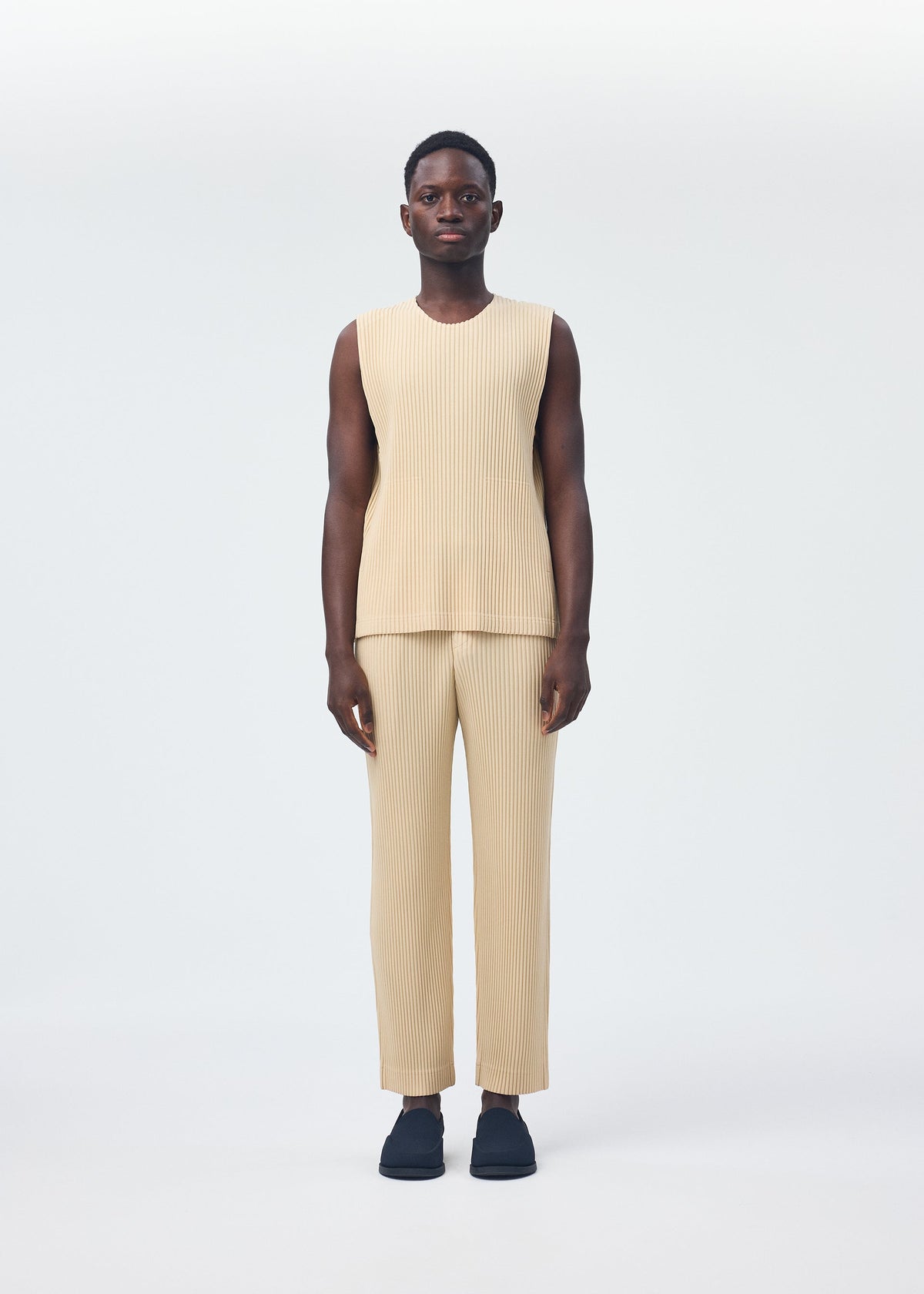 A person with short hair wears the TAILORED PLEATS 2 sleeveless beige set from HOMME PLISSÉ ISSEY MIYAKE—featuring a round-neck vest and ribbed quick-drying polyester pants—with dark slip-on shoes against a plain white background. view 6 of 8