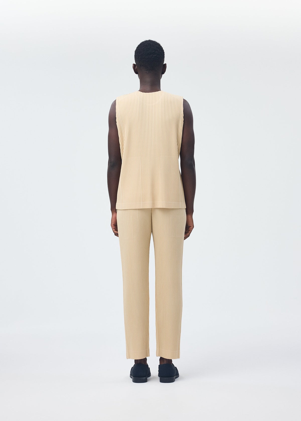 A person stands facing away from the camera, wearing the beige TAILORED PLEATS 2 set by HOMME PLISSÉ ISSEY MIYAKE—featuring a pleated round-neck vest and matching quick-dry polyester pants—with black shoes. view 8 of 8
