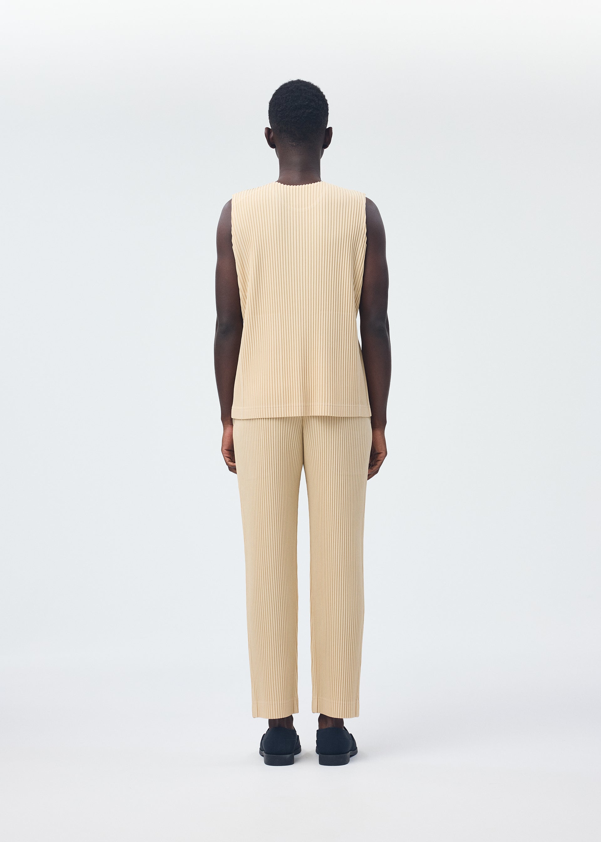 Zoomed view A person stands facing away from the camera, wearing the beige TAILORED PLEATS 2 set by HOMME PLISSÉ ISSEY MIYAKE—featuring a pleated round-neck vest and matching quick-dry polyester pants—with black shoes.