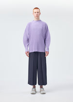 A person with short red hair wears a loose, light purple pleated MONTHLY COLORS : DECEMBER sweater and wide HOMME PLISSÉ ISSEY MIYAKE Japanese hakama pants with white shoes, standing against a plain white background.