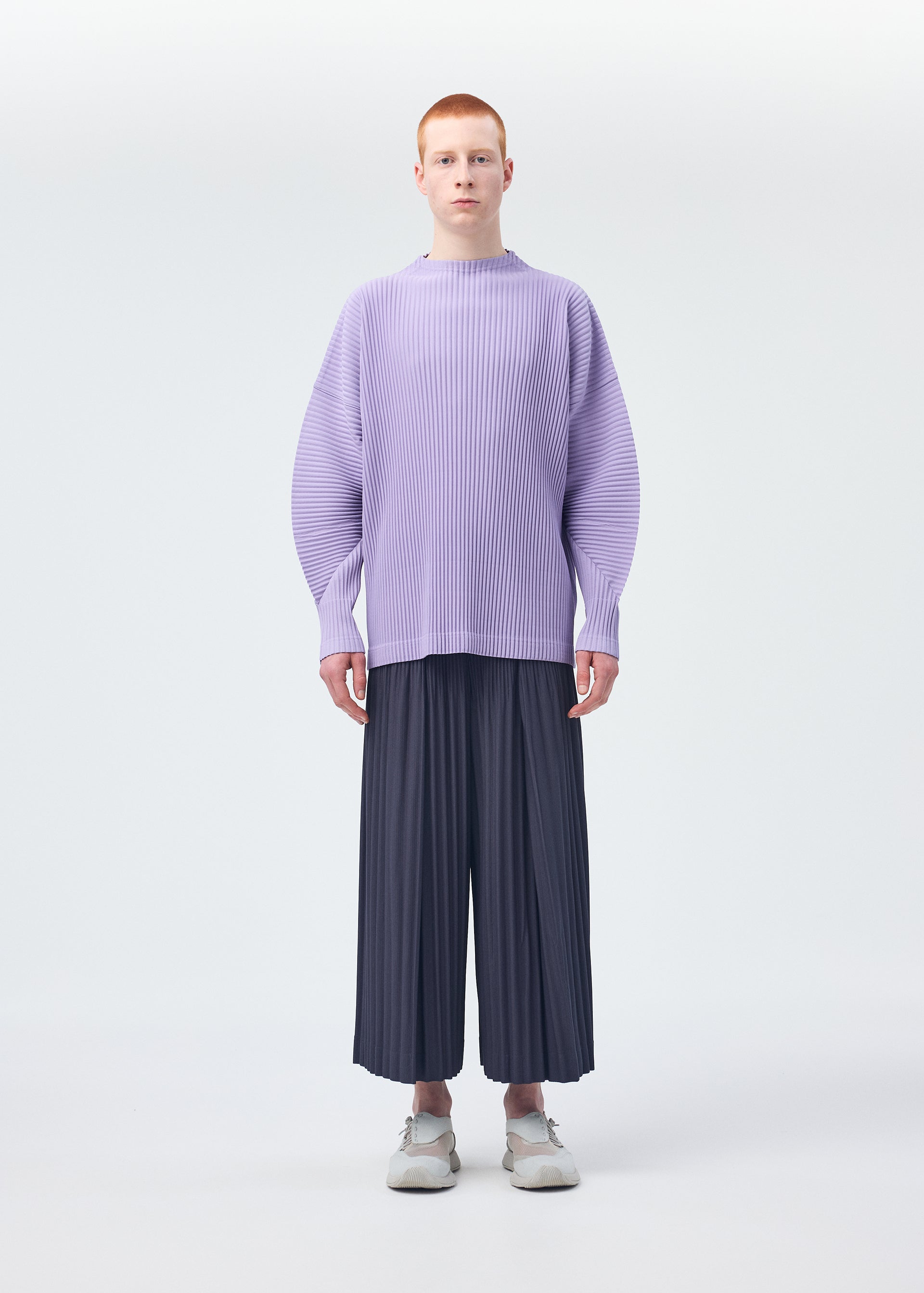 Zoomed view A person with short red hair wears a loose, light purple pleated MONTHLY COLORS : DECEMBER sweater and wide HOMME PLISSÉ ISSEY MIYAKE Japanese hakama pants with white shoes, standing against a plain white background.