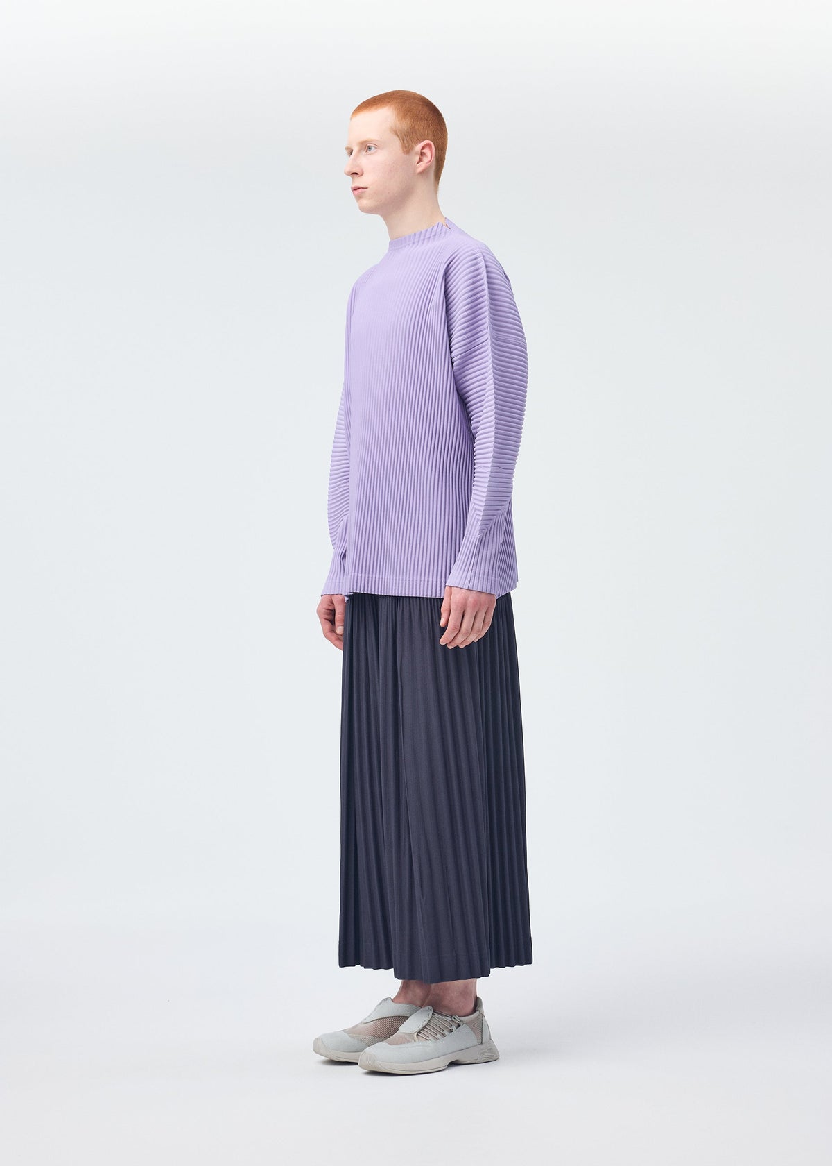 A person with short red hair stands in profile, wearing a loose, lavender long-sleeve top and HOMME PLISSÉ ISSEY MIYAKE MONTHLY COLORS : DECEMBER pleated dark wide pants, paired with white sneakers in a minimal white studio. view 7 of 8