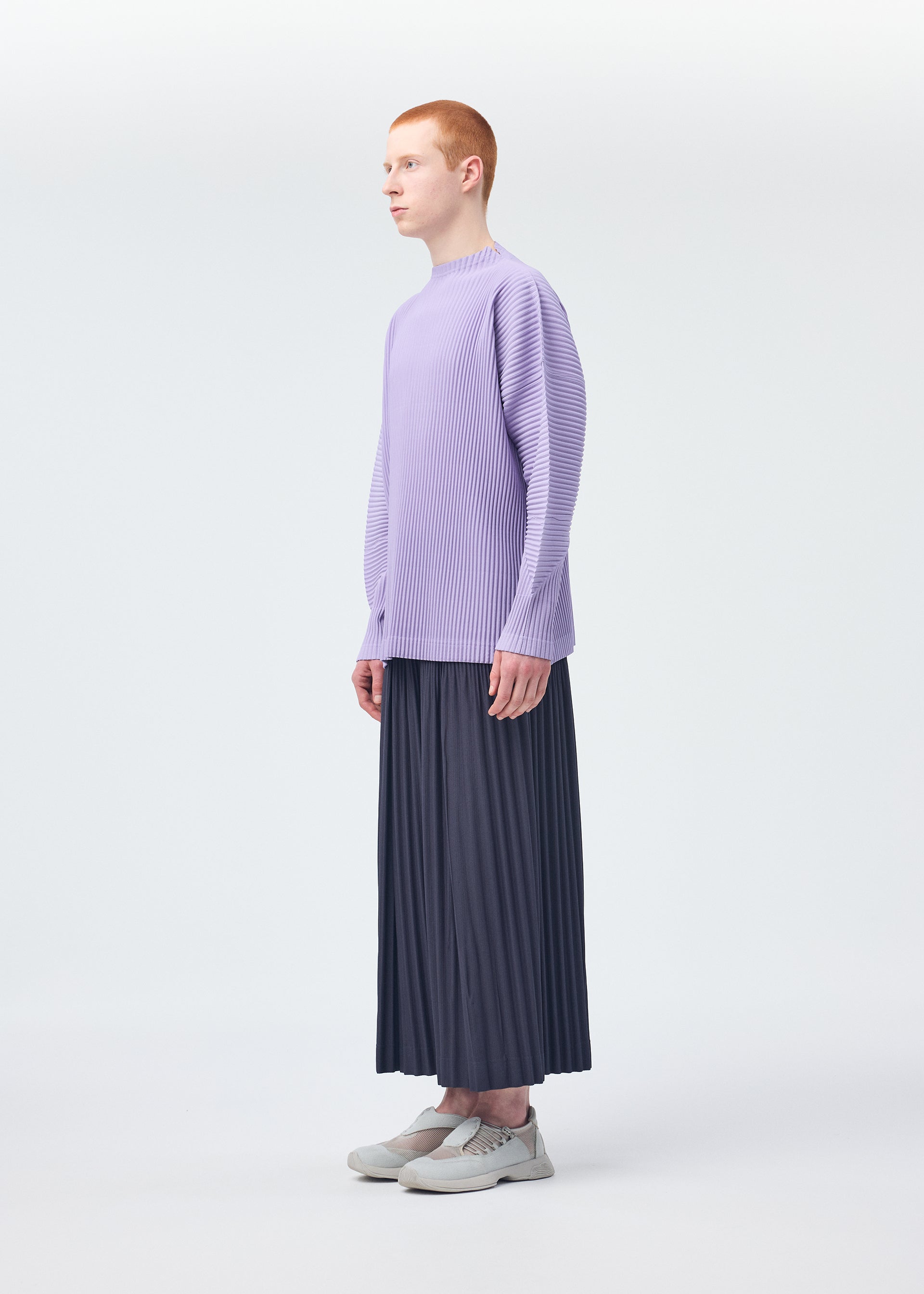 Zoomed view A person with short red hair stands in profile, wearing a loose, lavender long-sleeve top and HOMME PLISSÉ ISSEY MIYAKE MONTHLY COLORS : DECEMBER pleated dark wide pants, paired with white sneakers in a minimal white studio.