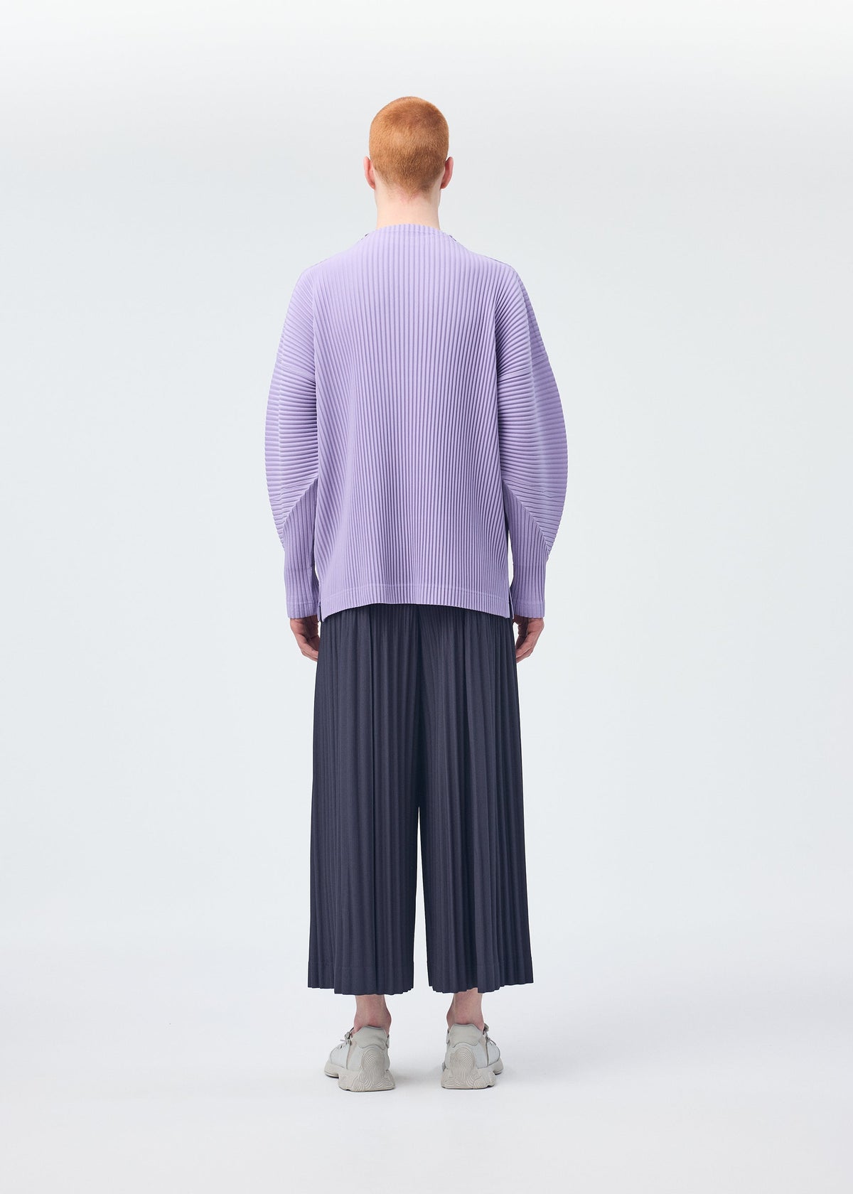 A person with short red hair faces away, wearing HOMME PLISSÉ ISSEY MIYAKE's MONTHLY COLORS : DECEMBER—a loose lavender pleated top and wide dark hakama pants—paired with white sneakers against a plain light background. view 8 of 8