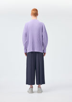 A person with short red hair faces away, wearing HOMME PLISSÉ ISSEY MIYAKE's MONTHLY COLORS : DECEMBER—a loose lavender pleated top and wide dark hakama pants—paired with white sneakers against a plain light background.