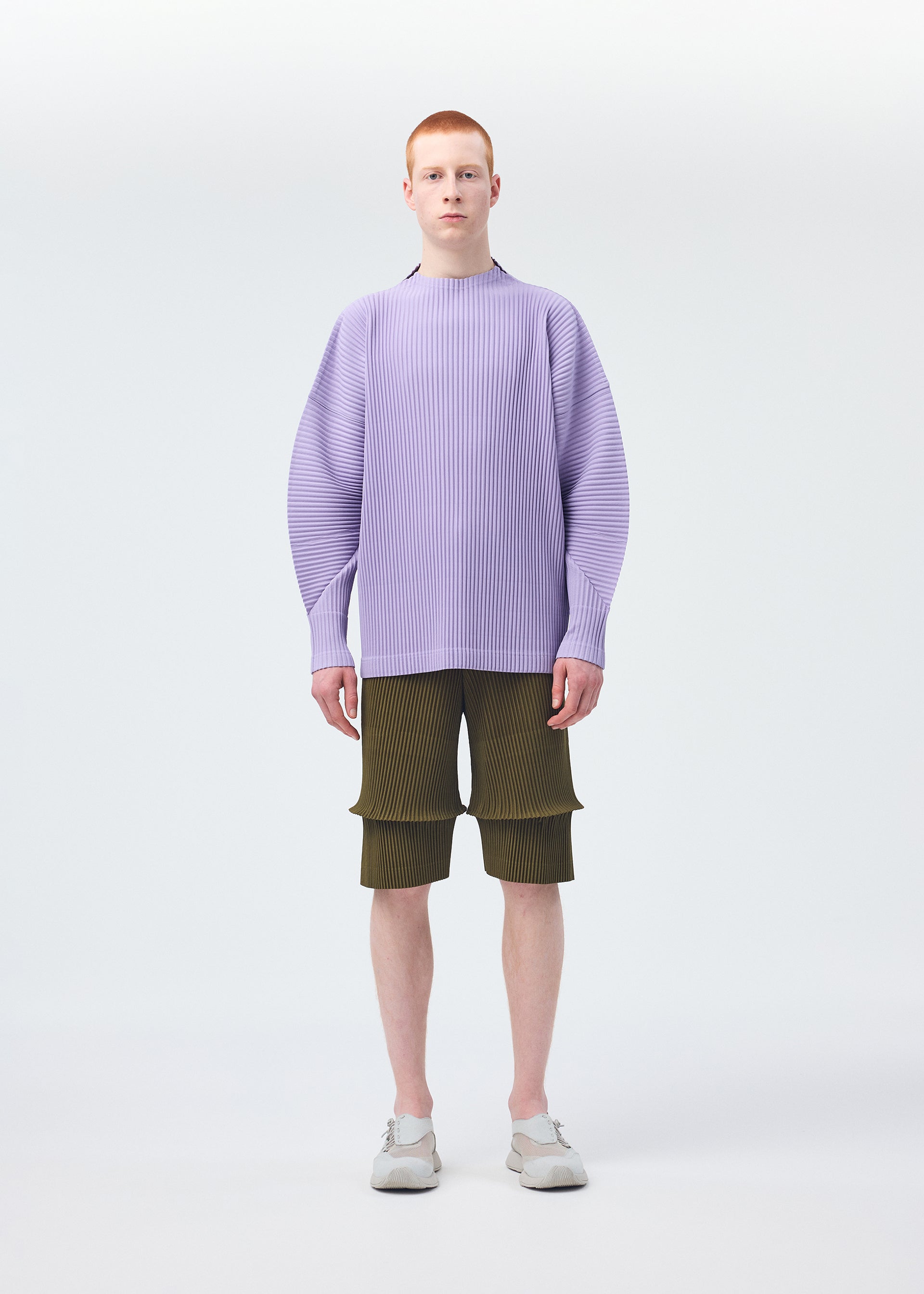 Zoomed view A person with short red hair faces forward in a loose light purple ribbed sweater, olive green HOMME PLISSÉ ISSEY MIYAKE PLEATS BOTTOMS 2 shorts with pockets and panels, and light gray slip-on shoes against a plain white background.