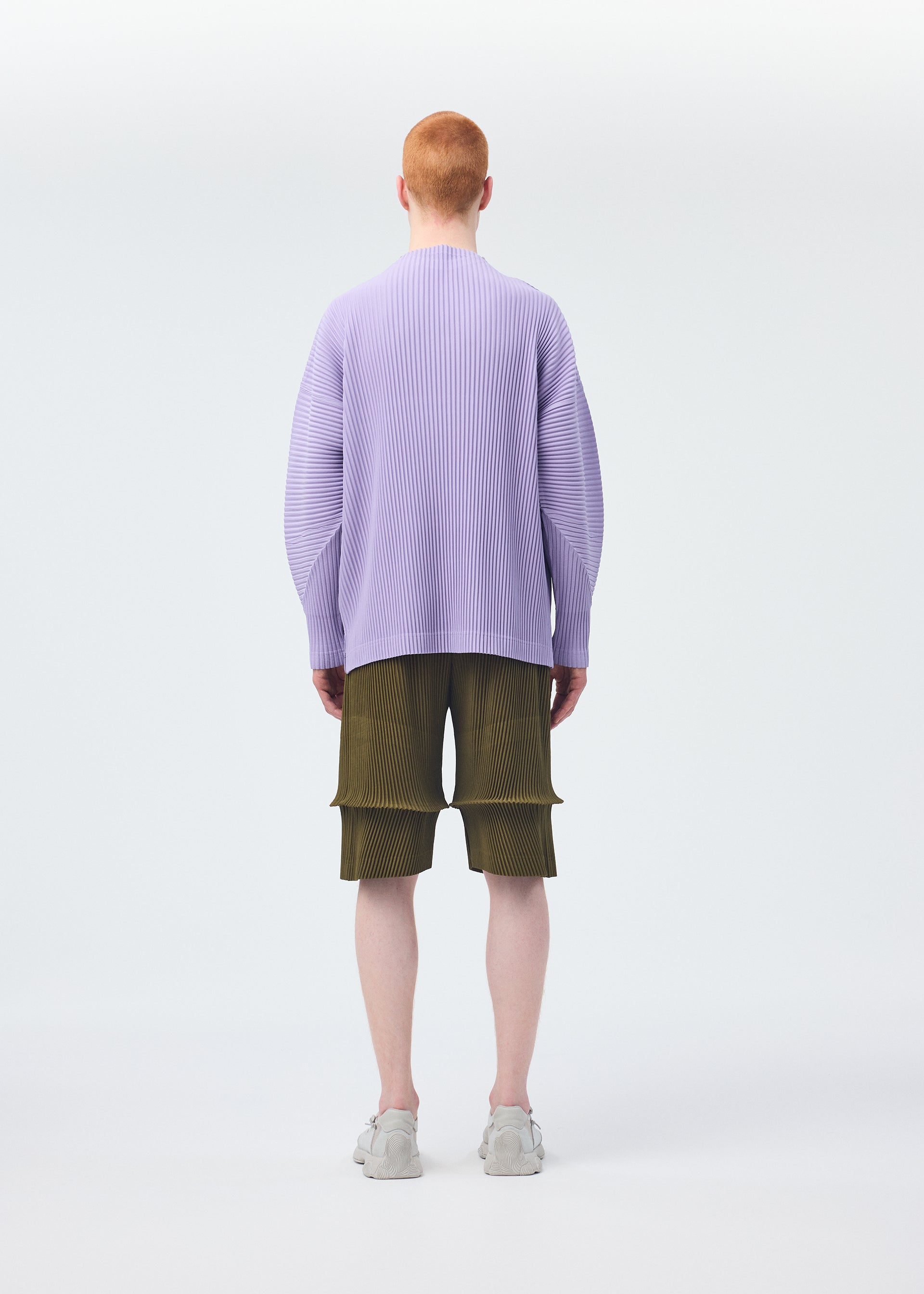 Zoomed view A person with short red hair, seen from behind against a white background, wears a loose lavender sweater, white sneakers, and HOMME PLISSÉ ISSEY MIYAKE PLEATS BOTTOMS 2 shorts with pockets.