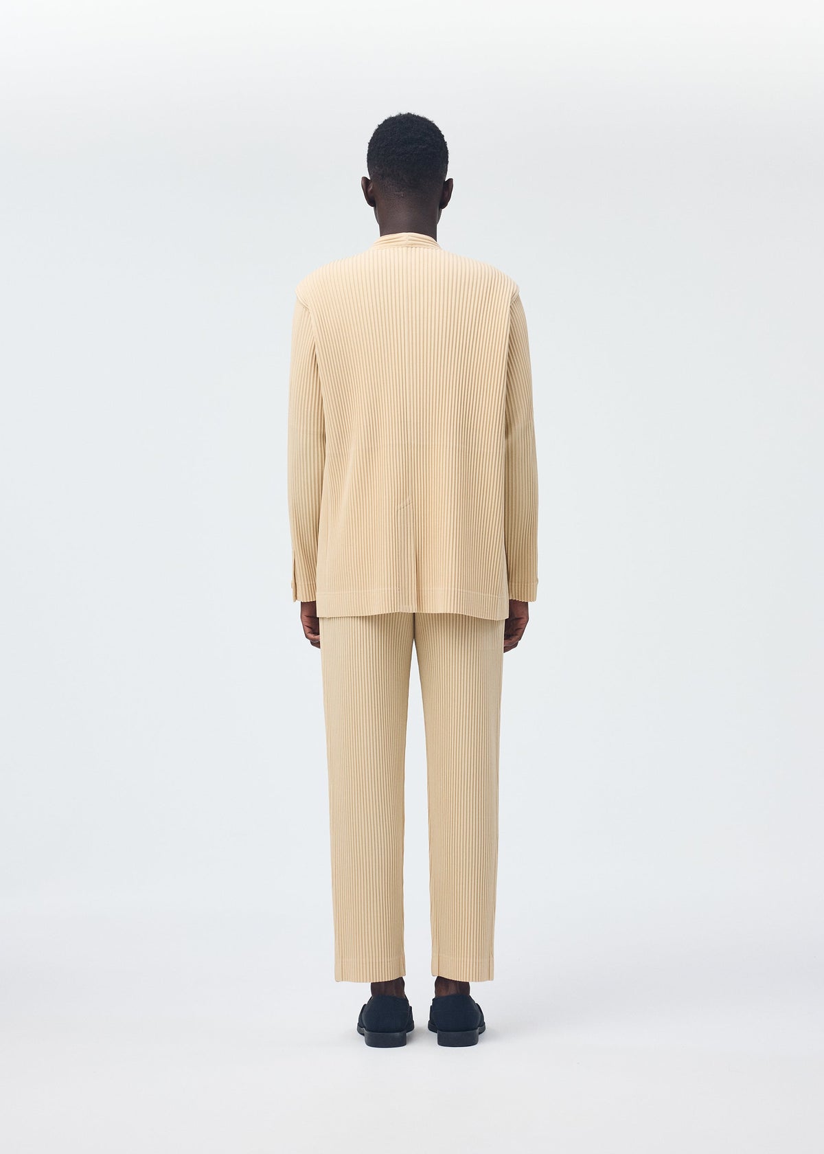 A person stands facing away, wearing the HOMME PLISSÉ ISSEY MIYAKE TAILORED PLEATS 2 set—a beige, pleated long-sleeve top and straight pants in quick-drying polyester—styled with dark shoes against a plain white background. view 7 of 7