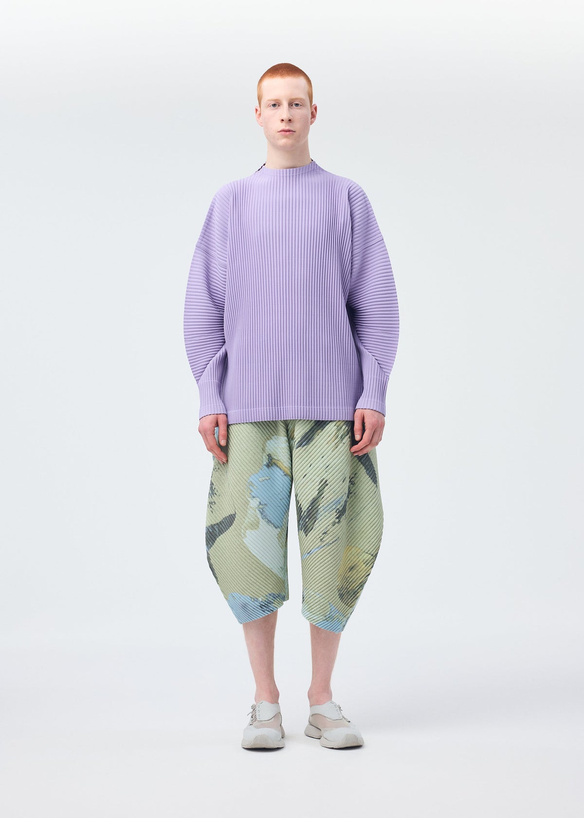 A person faces forward in a loose, light purple ribbed sweater and HOMME PLISSÉ ISSEY MIYAKE PAINTED STONE baggy green pants with blue and gray shapes, paired with white shoes against a plain white background. view 6 of 8