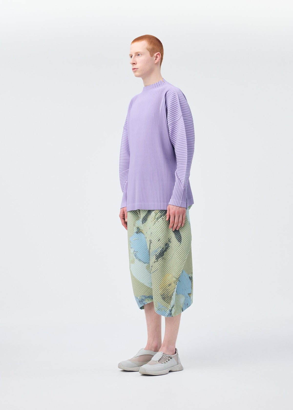 A person with short red hair stands sideways in a studio, wearing HOMME PLISSÉ ISSEY MIYAKE’s PAINTED STONE pattern-painted shorts, a lavender ribbed top, and light gray slip-on shoes against a white background. view 7 of 8