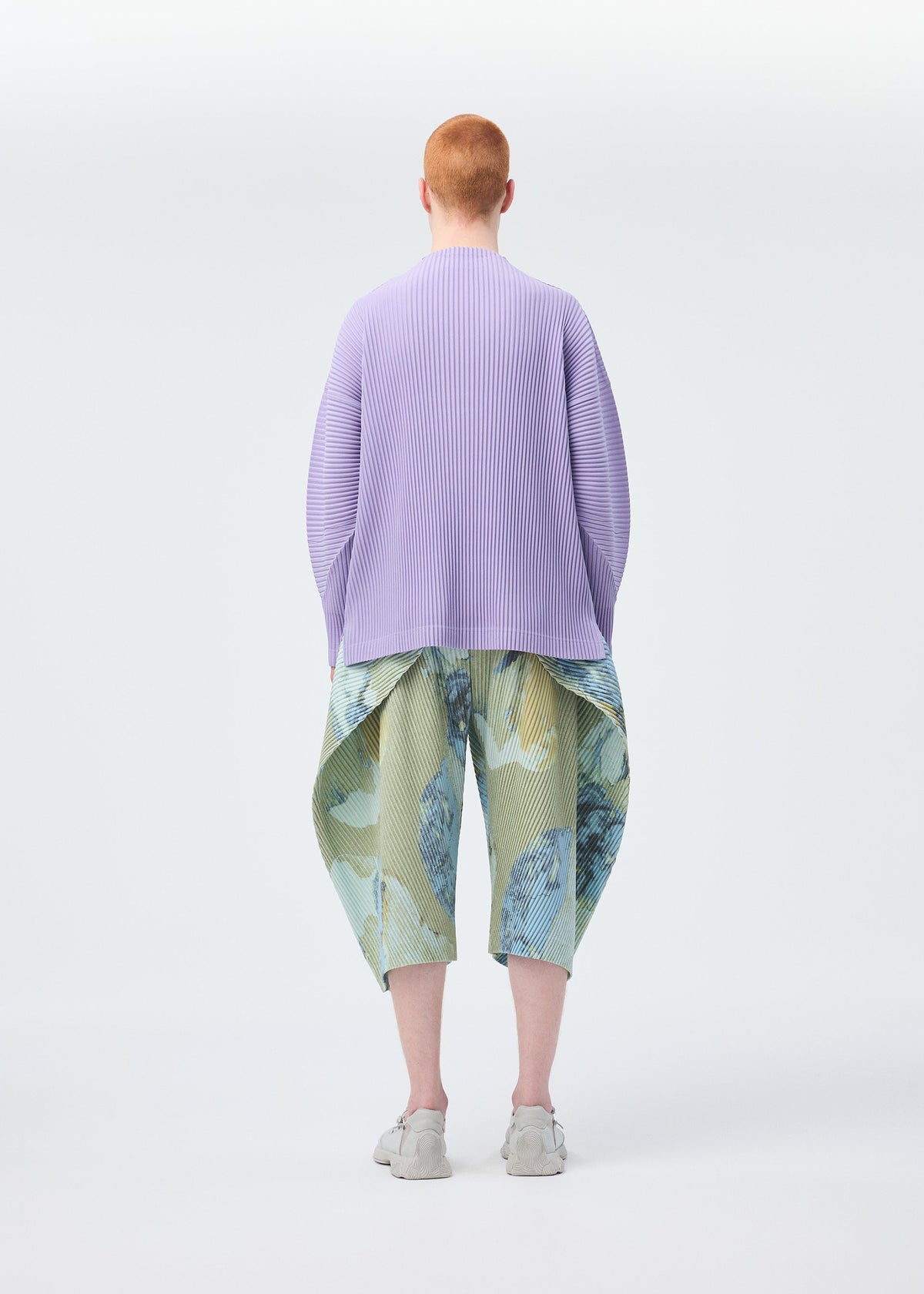A person with short red hair, seen from behind, wears HOMME PLISSÉ ISSEY MIYAKE's PAINTED STONE pastel green and blue textured pants, a lavender long-sleeve top, and light gray shoes against a plain white backdrop. view 8 of 8