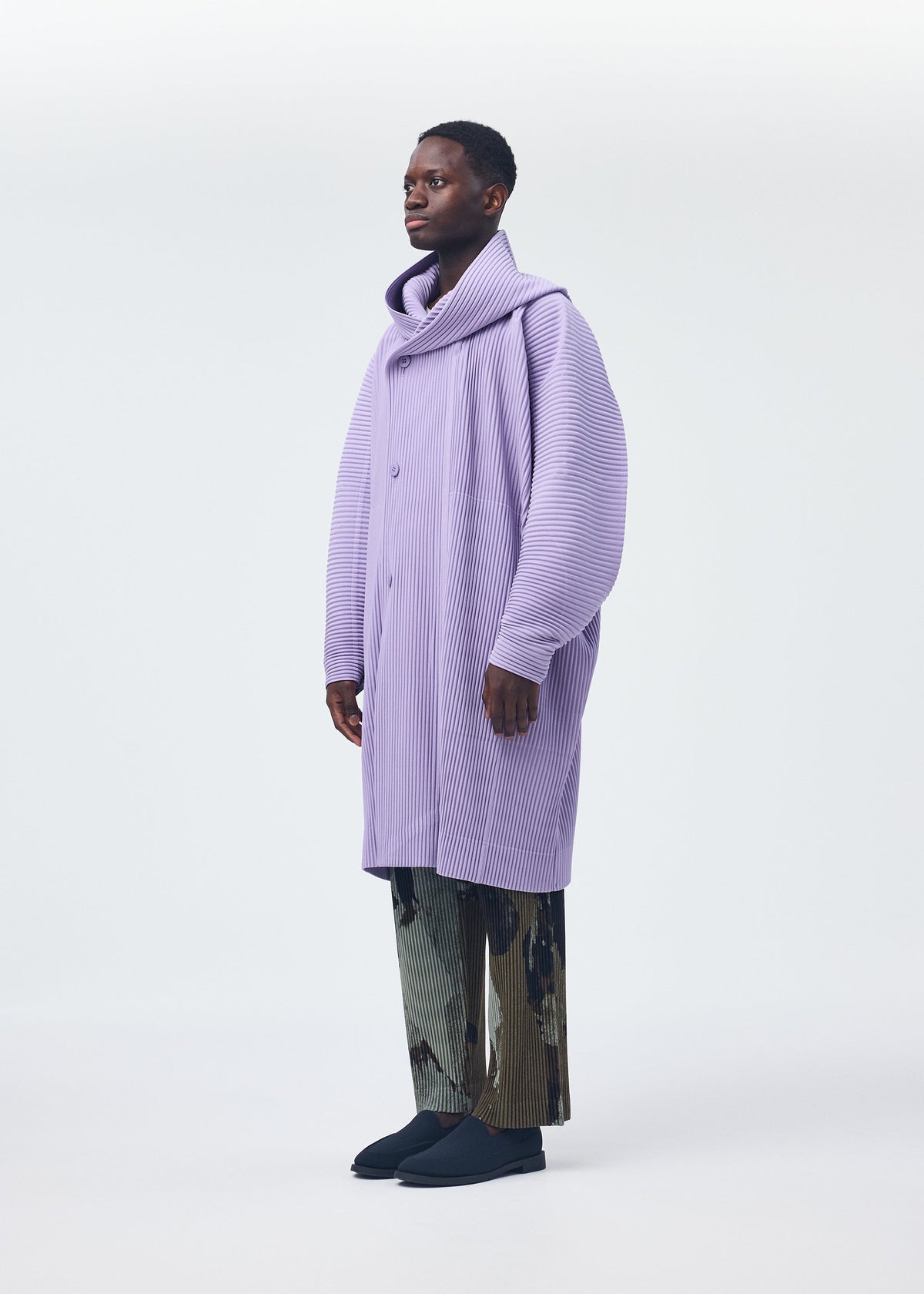 A person poses against a plain background, wearing HOMME PLISSÉ ISSEY MIYAKE’s PAINTED STONE: an oversized, light purple textured coat with large lapels and straight, subtly stone-patterned pants, paired with dark shoes for a modern look. view 7 of 8