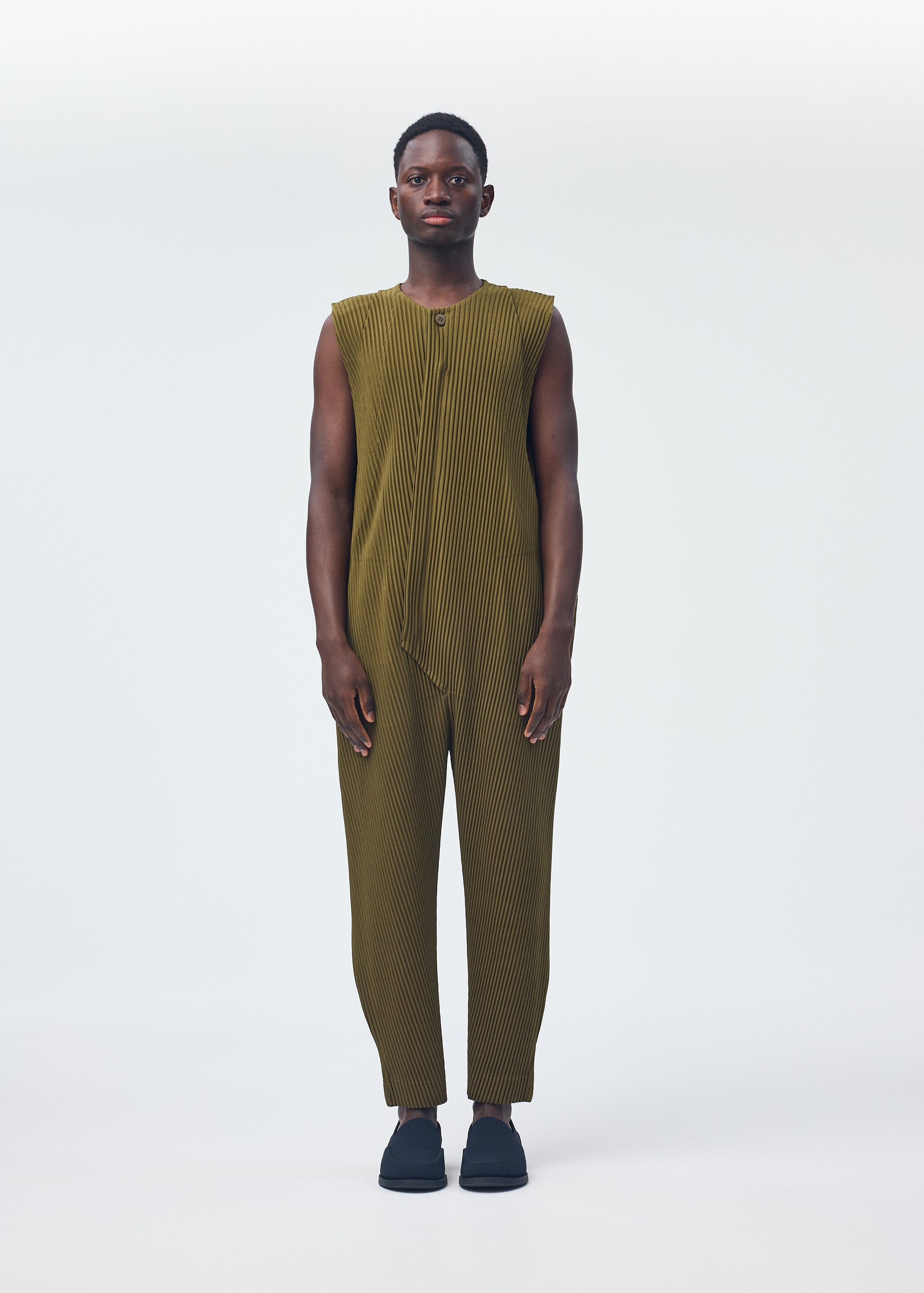 Zoomed view A person faces forward, wearing HOMME PLISSÉ ISSEY MIYAKE PLEATS BOTTOMS 2—a comfortable, sleeveless olive-green jumpsuit with a zipper fly—paired with black slip-on shoes, set against a plain light background.