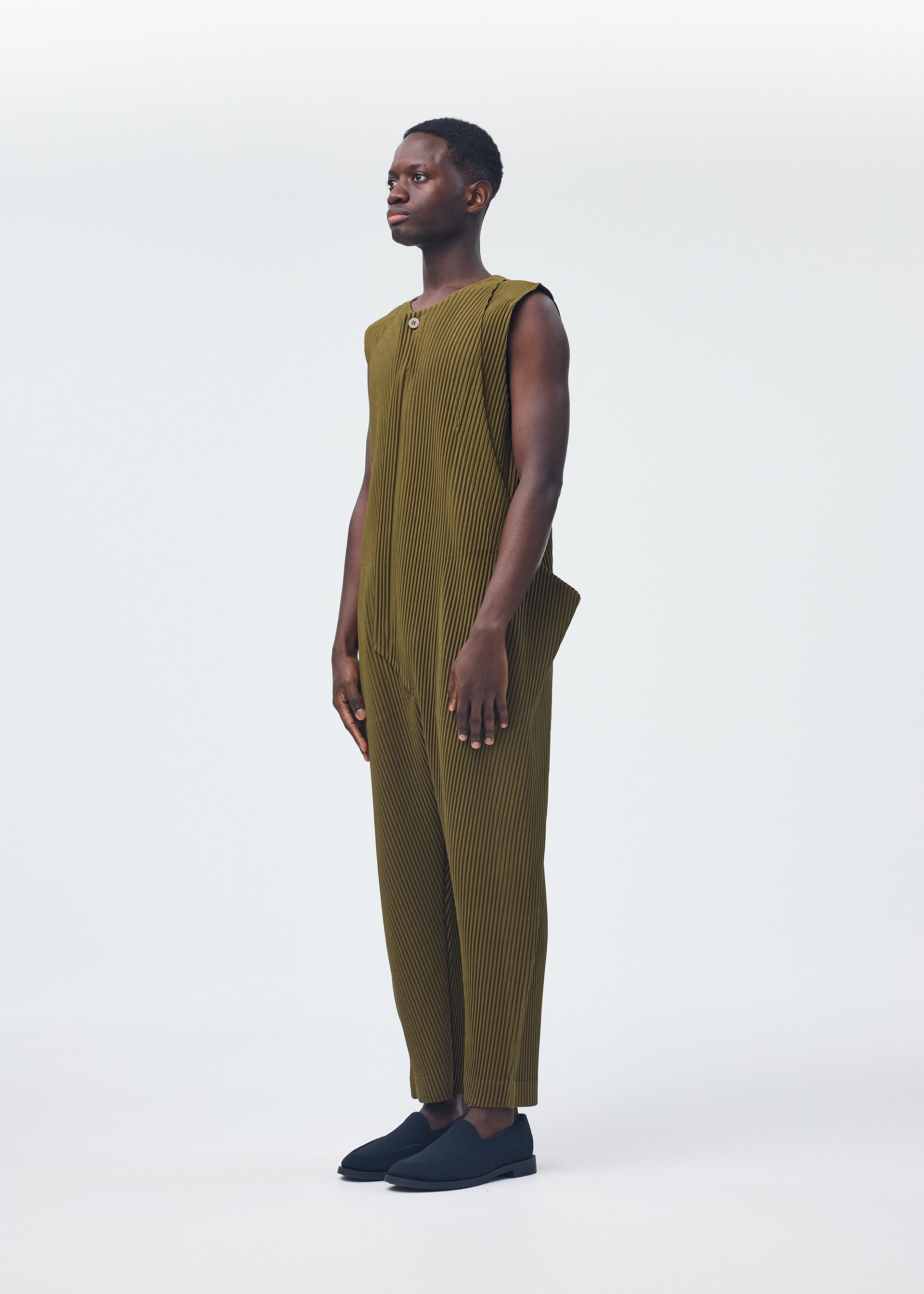 Zoomed view A person faces left, wearing the HOMME PLISSÉ ISSEY MIYAKE PLEATS BOTTOMS 2—an olive green, sleeveless pleated jumpsuit with cropped legs and a zipper fly—paired with black slip-on shoes against a plain white background.