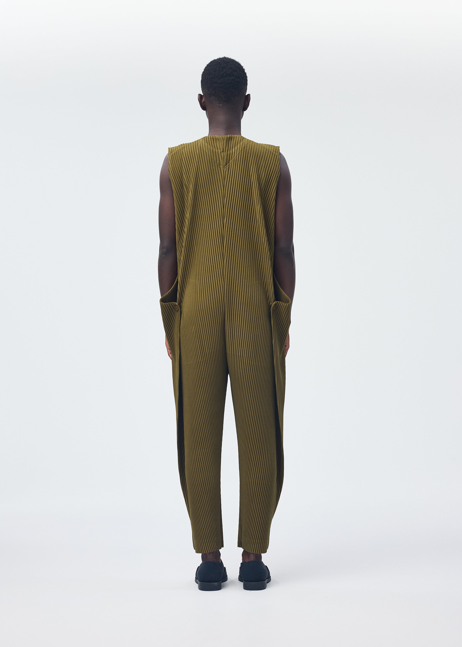 Zoomed view A person faces away, wearing HOMME PLISSÉ ISSEY MIYAKE’s PLEATS BOTTOMS 2 in olive-green with vertical pleats and a zipper fly, paired with black shoes against a plain light background.