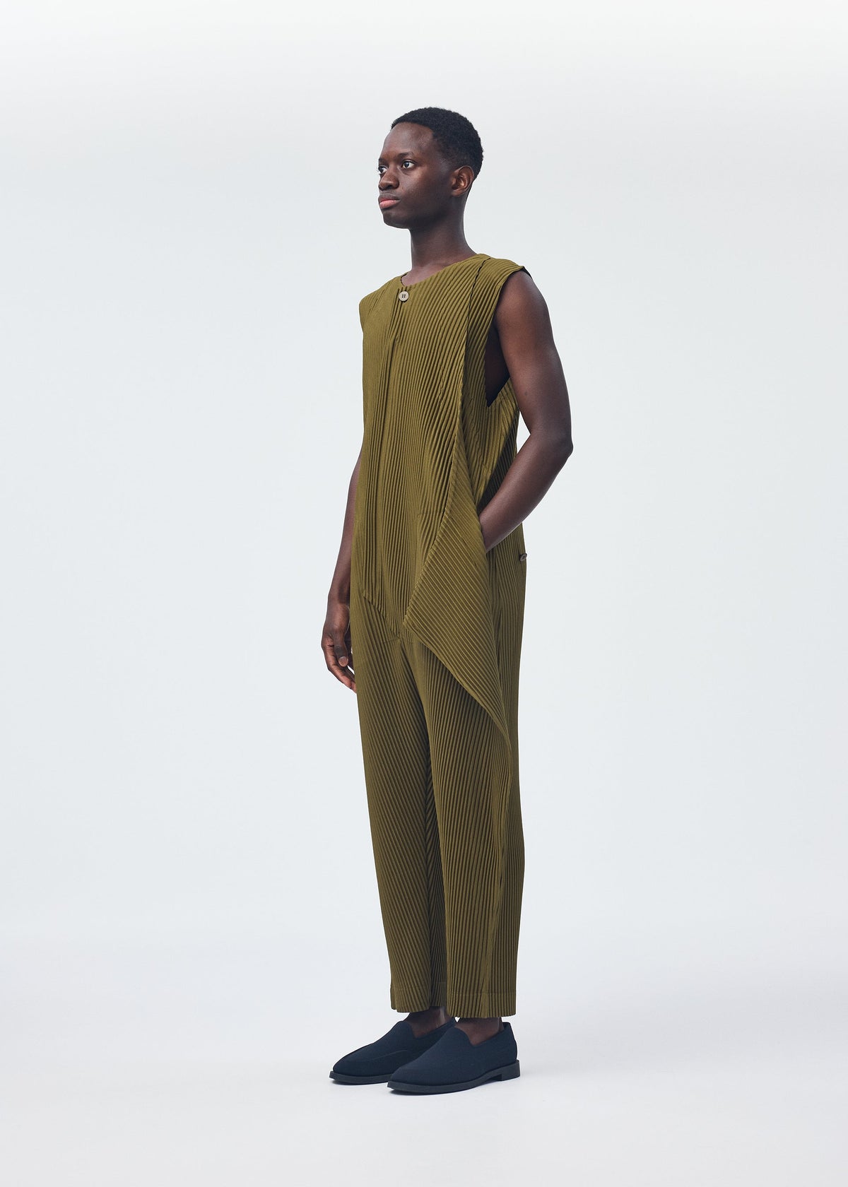 A person stands against a white background wearing HOMME PLISSÉ ISSEY MIYAKE PLEATS BOTTOMS 2—comfortable, olive-green, wide-leg pleated pants with side draping and a zipper fly—paired with black slip-on shoes. view 9 of 9
