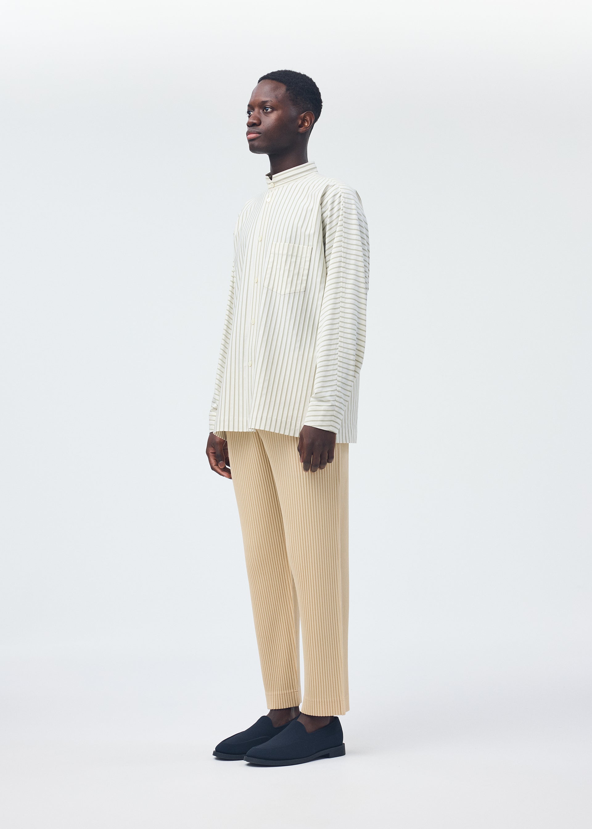 Zoomed view A man wears the HOMME PLISSÉ ISSEY MIYAKE STRIPE JERSEY SHIRT in cream and white stripes, with beige striped pants and black slip-ons, standing against a white background and facing slightly left with a neutral expression.