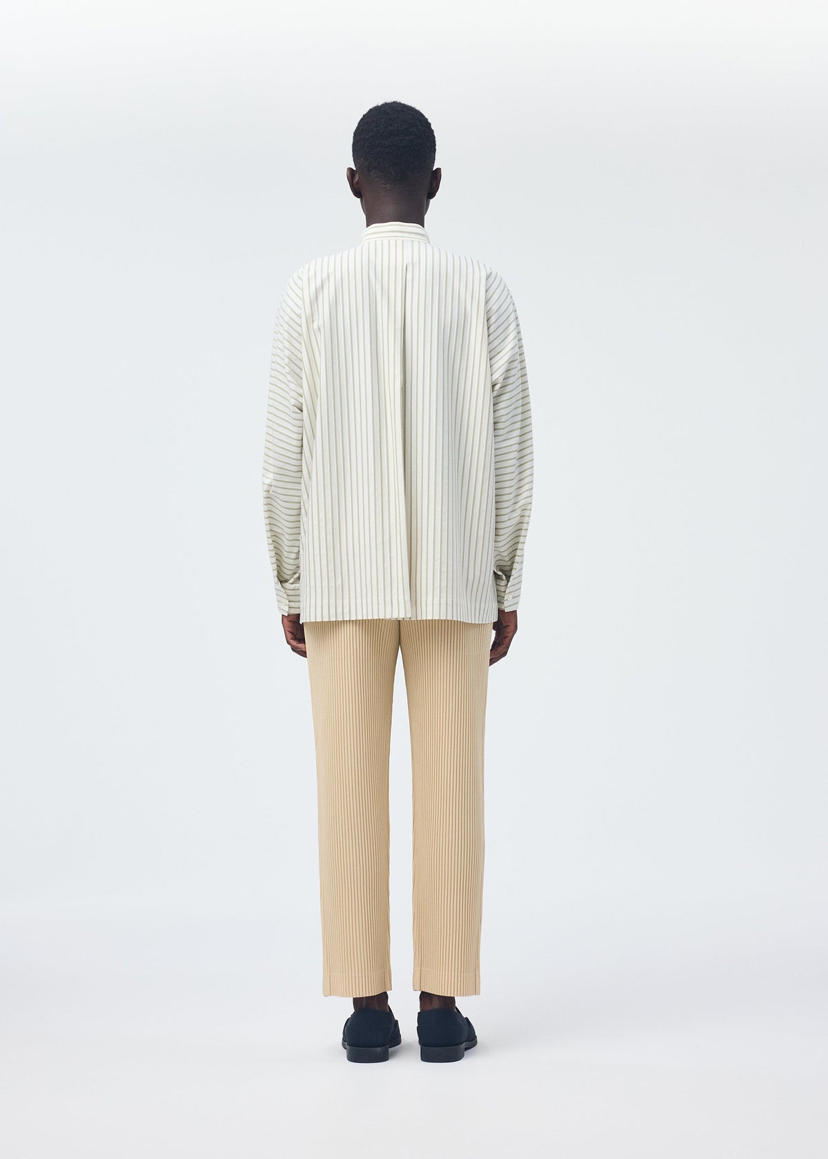 A person faces away from the camera in a STRIPE JERSEY SHIRT by HOMME PLISSÉ ISSEY MIYAKE, featuring a loose fit and stripes, paired with beige pleated pants and black shoes against a plain white background. view 8 of 8