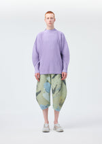 A person stands facing forward against a plain light background, dressed in HOMME PLISSÉ ISSEY MIYAKE’s MONTHLY COLORS : DECEMBER—loose lavender ribbed long-sleeved top, wide abstract green and blue knee-length shorts, and white shoes.