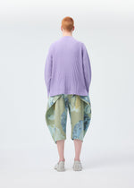 A person with short red hair stands facing away, wearing HOMME PLISSÉ ISSEY MIYAKE's MONTHLY COLORS : DECEMBER collection—a loose, ribbed lavender mock neck top and wide blue-green patterned shorts—against a plain light background.