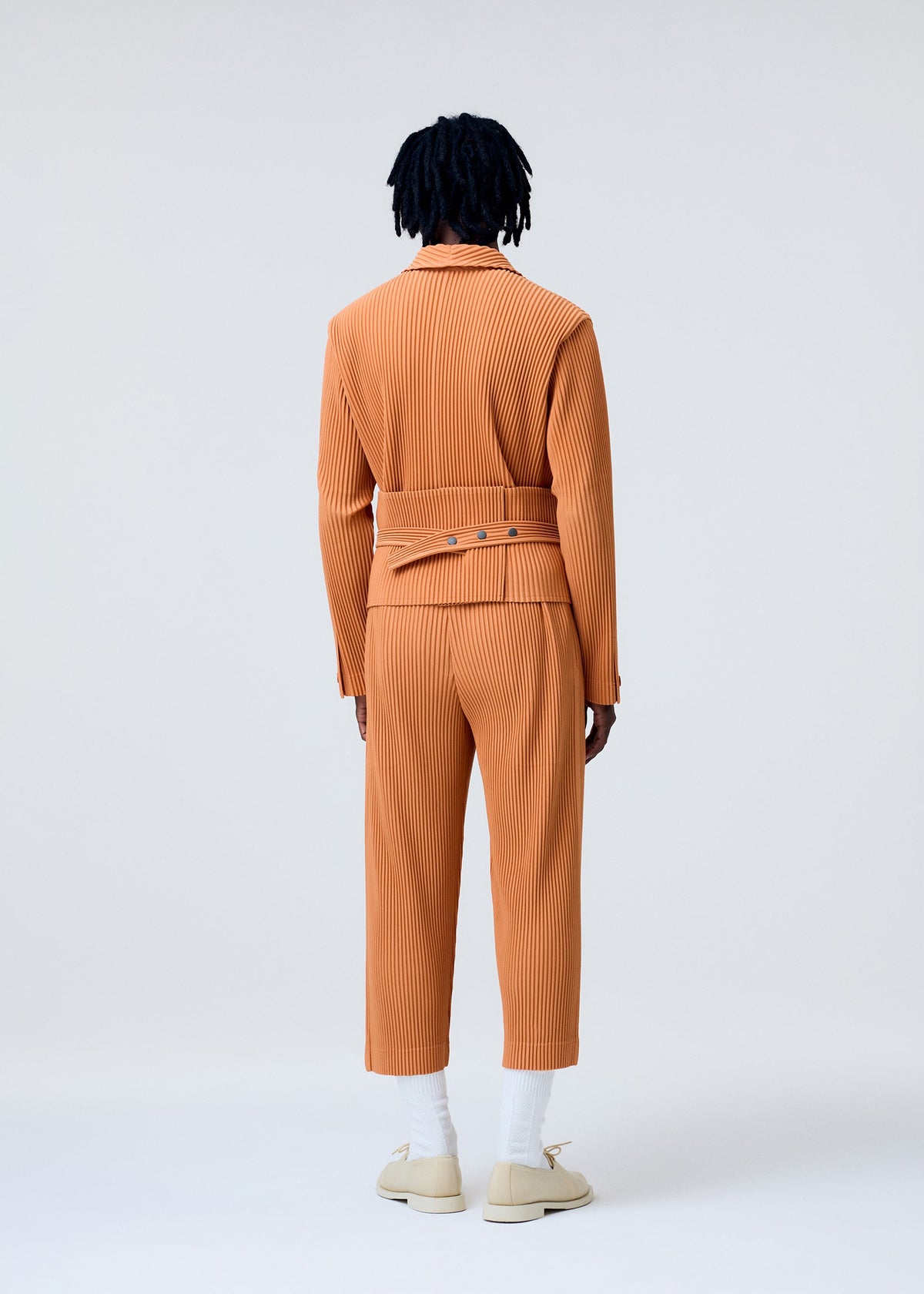 A person with dark, short dreadlocks stands facing away, wearing the orange PLEATS BAND set by HOMME PLISSÉ ISSEY MIYAKE, with white socks and cream shoes against a plain light gray background. view 6 of 8