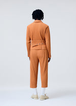 A person with dark, short dreadlocks stands facing away, wearing the orange PLEATS BAND set by HOMME PLISSÉ ISSEY MIYAKE, with white socks and cream shoes against a plain light gray background.