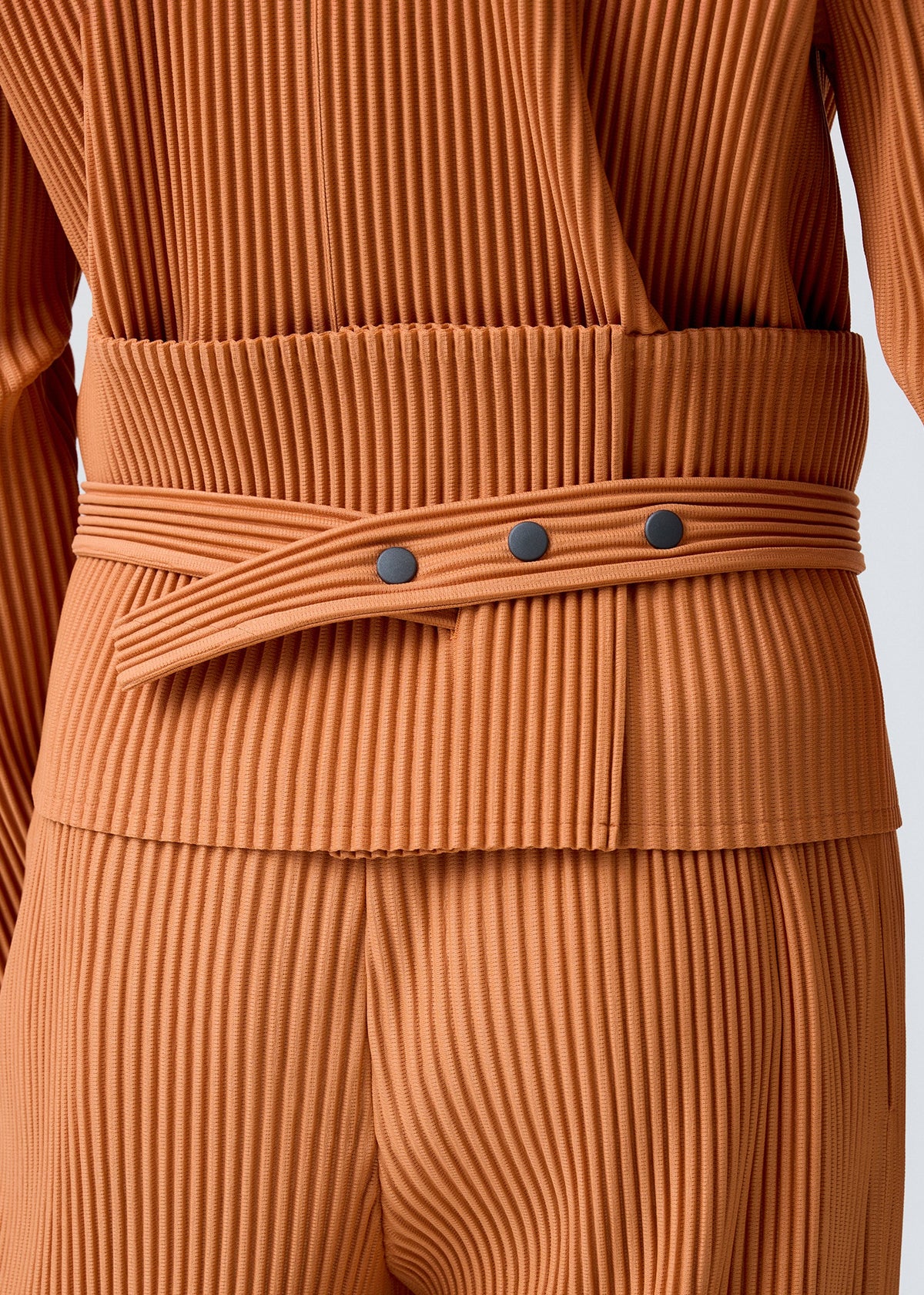 Close-up of a person wearing the PLEATS BAND by HOMME PLISSÉ ISSEY MIYAKE—a rust dress crafted from tube-shaped fabric with vertical ridges, cinched at the waist by a wide matching belt with three black buttons and overlapping ends. view 7 of 8