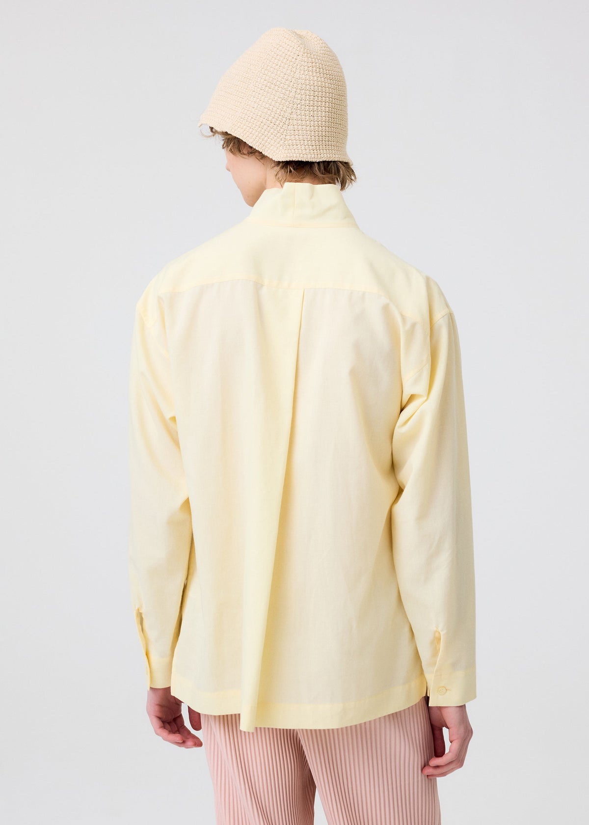 A person wearing a pale yellow long-sleeve shirt, pink striped pants, and the HOMME PLISSÉ ISSEY MIYAKE CROCHET HAT stands facing away from the camera against a plain light background. view 5 of 5