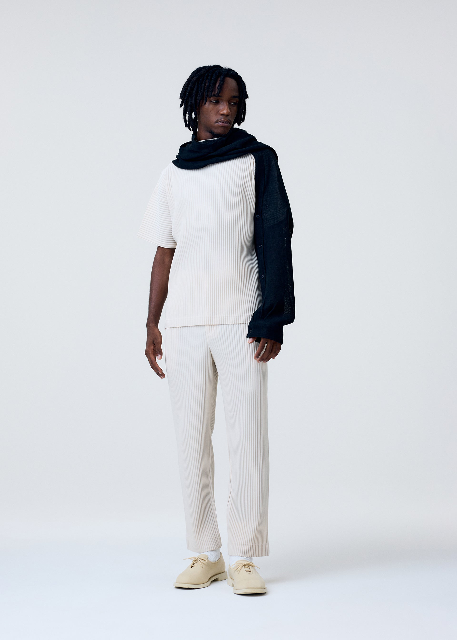 Zoomed view A person with short dreadlocks and a neutral expression stands against a plain light background, wearing the HOMME PLISSÉ ISSEY MIYAKE SLEEVED STOLE draped over one shoulder, paired with cream ribbed top, matching pants, and cream shoes.