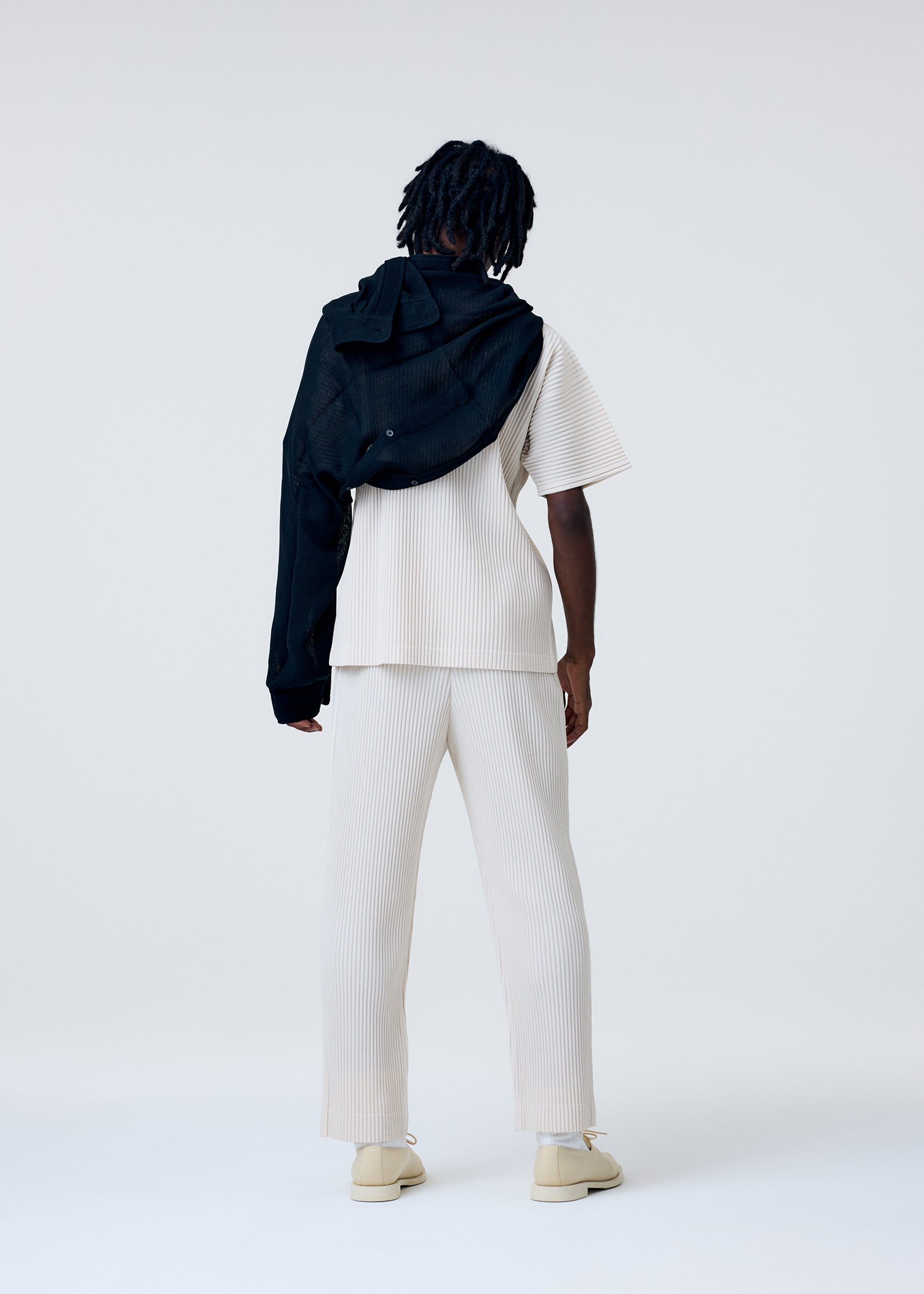 Zoomed view A person faces away wearing a cream ribbed short-sleeve top and pants. A black HOMME PLISSÉ ISSEY MIYAKE SLEEVED STOLE is draped over one shoulder. The background is plain and light-colored.