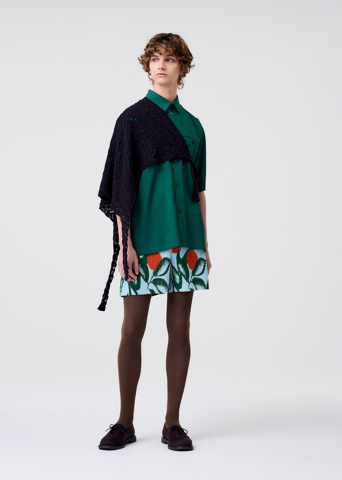 Wearing HOMME PLISSÉ ISSEY MIYAKE’s BRANCHES & LEAVES skirt, a person with short wavy hair pairs it with a green shirt, black lace shawl, brown tights, dark shoes, and a textured scarf against a plain light background. view 5 of 7