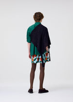 A person faces away wearing HOMME PLISSÉ ISSEY MIYAKE's BRANCHES & LEAVES, styled with a green shirt, a floral orange-green skirt, brown tights, black shoes, and a knitted scarf. The background is plain and light-colored.