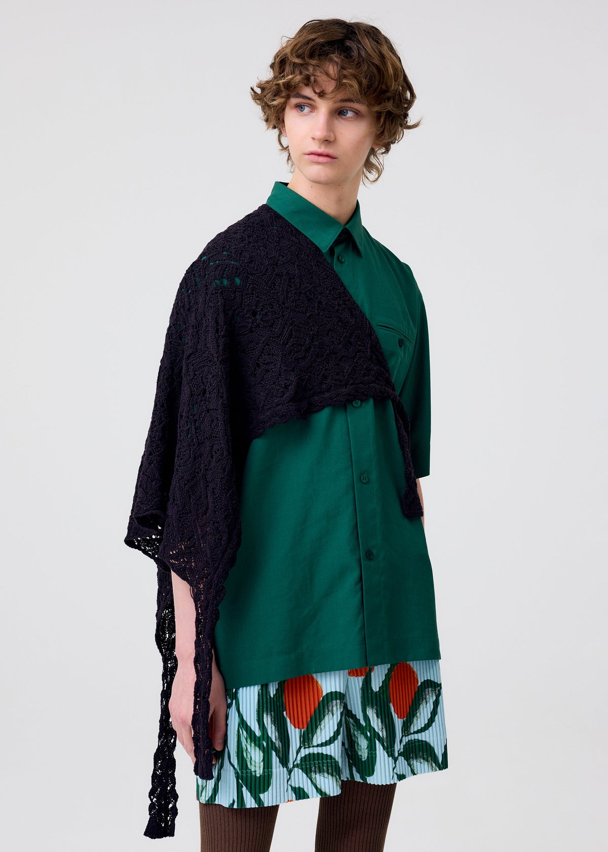 A person with short, curly hair wears the HOMME PLISSÉ ISSEY MIYAKE BRANCHES & LEAVES skirt featuring colorful leaf and flower patterns, paired with a green button-up shirt and black lace shawl, looking to the side against a plain background. view 7 of 7