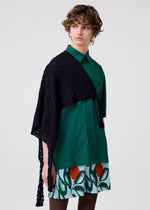 A person with short, curly hair wears the HOMME PLISSÉ ISSEY MIYAKE BRANCHES & LEAVES skirt featuring colorful leaf and flower patterns, paired with a green button-up shirt and black lace shawl, looking to the side against a plain background.