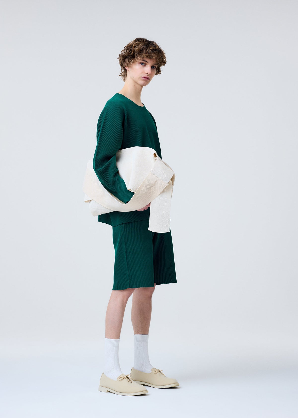 A person with curly hair wears a loose green long-sleeve shirt, matching knee-length shorts, white socks, beige shoes, and carries the HOMME PLISSÉ ISSEY MIYAKE WASHI FLAP BAG while holding a folded cream garment against a plain light background. view 5 of 6