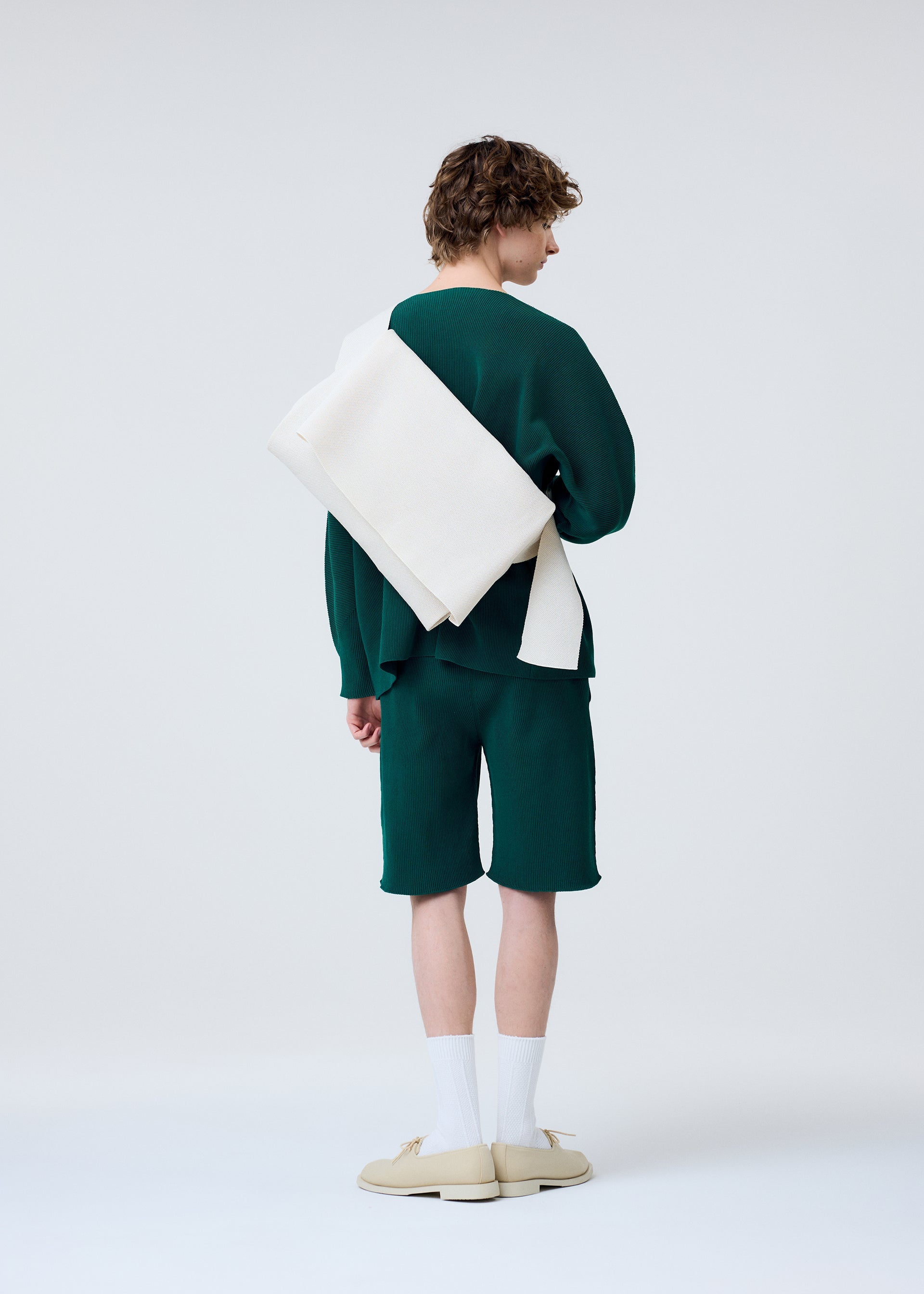 Zoomed view A person with curly hair, dressed in a green long-sleeve top, matching shorts, white socks, and beige shoes, stands facing away. They carry the HOMME PLISSÉ ISSEY MIYAKE WASHI FLAP BAG on their shoulder against a plain background.