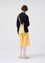 A person with short hair faces away, wearing a dark long-sleeve top, yellow pleated culottes, white socks, beige shoes, and the HOMME PLISSÉ ISSEY MIYAKE DRAWSTRING PLEATS BAG in yellow across the body against a plain light background.