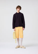 A person with wavy hair stands against a plain light background wearing a loose dark long-sleeve top, light yellow pleated knee-length shorts, white tights, cream shoes, and holding the HOMME PLISSÉ ISSEY MIYAKE DRAWSTRING PLEATS BAG.