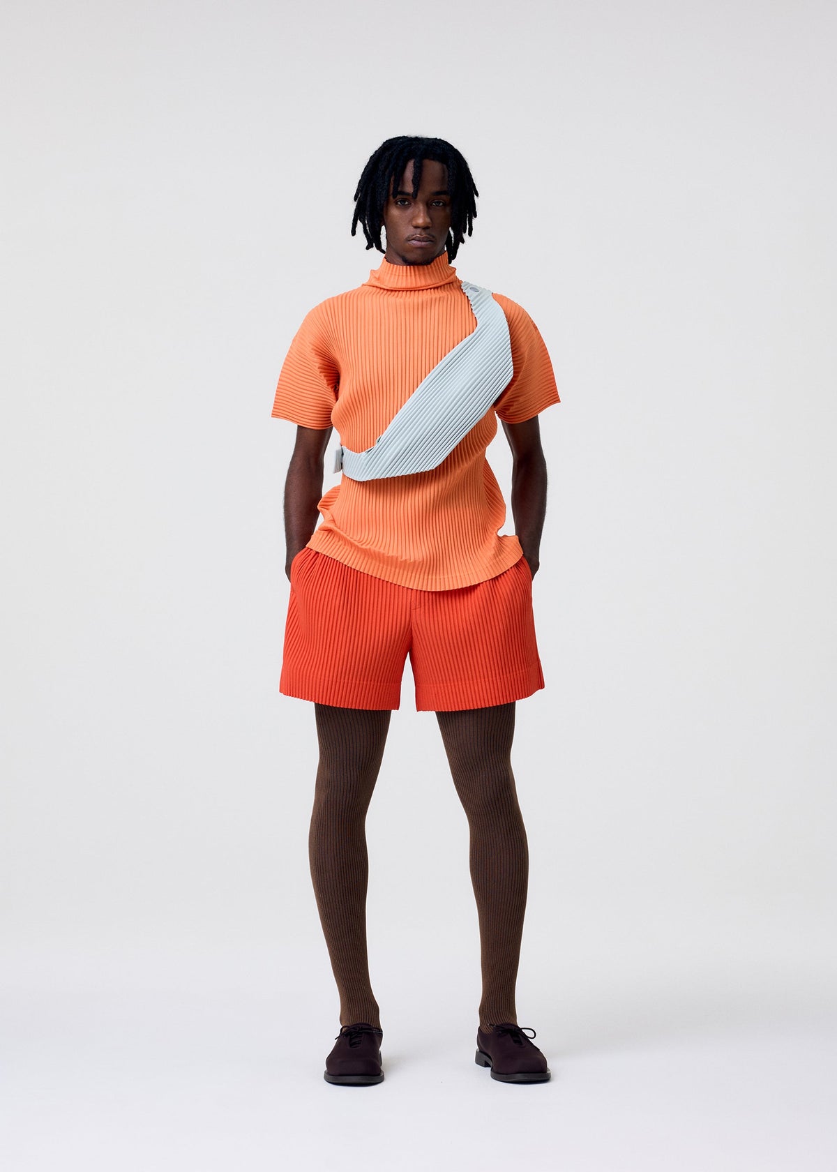 A person stands against a plain background wearing an orange short-sleeve top, matching shorts, brown tights, dark shoes, and carries the HOMME PLISSÉ ISSEY MIYAKE VERSATILE CROSSBODY in light blue. Hands are in pockets and expression is neutral. view 7 of 8