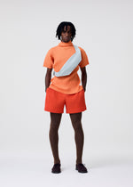 A person stands against a plain background wearing an orange short-sleeve top, matching shorts, brown tights, dark shoes, and carries the HOMME PLISSÉ ISSEY MIYAKE VERSATILE CROSSBODY in light blue. Hands are in pockets and expression is neutral.