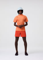 A person stands against a plain background wearing a light blue hat, an orange ribbed short-sleeve top, red-orange shorts, brown textured tights, dark shoes, and carries the HOMME PLISSÉ ISSEY MIYAKE VERSATILE CROSSBODY, hands clasped at their waist.