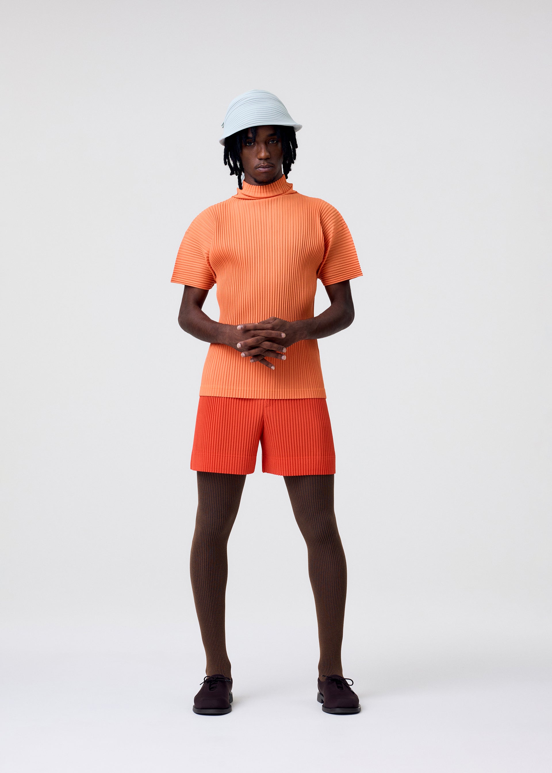Zoomed view A person stands against a plain background wearing a light blue hat, an orange ribbed short-sleeve top, red-orange shorts, brown textured tights, dark shoes, and carries the HOMME PLISSÉ ISSEY MIYAKE VERSATILE CROSSBODY, hands clasped at their waist.