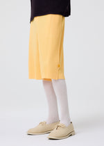 A person wearing yellow pleated knee-length shorts, beige shoes, a dark top, and HOMME PLISSÉ ISSEY MIYAKE WASHI BLEND SOCKS stands against a plain white background.