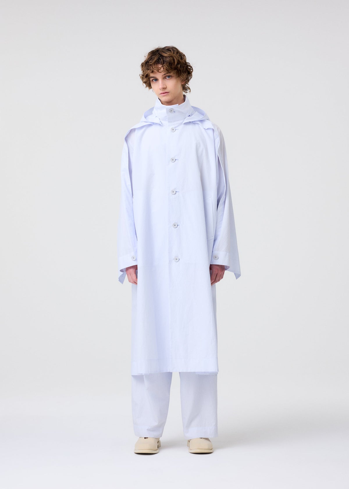 A person with short curly hair wears the FRESHEN hooded coat and matching wide-legged pants by HOMME PLISSÉ ISSEY MIYAKE, styled with beige shoes against a plain background. view 7 of 9