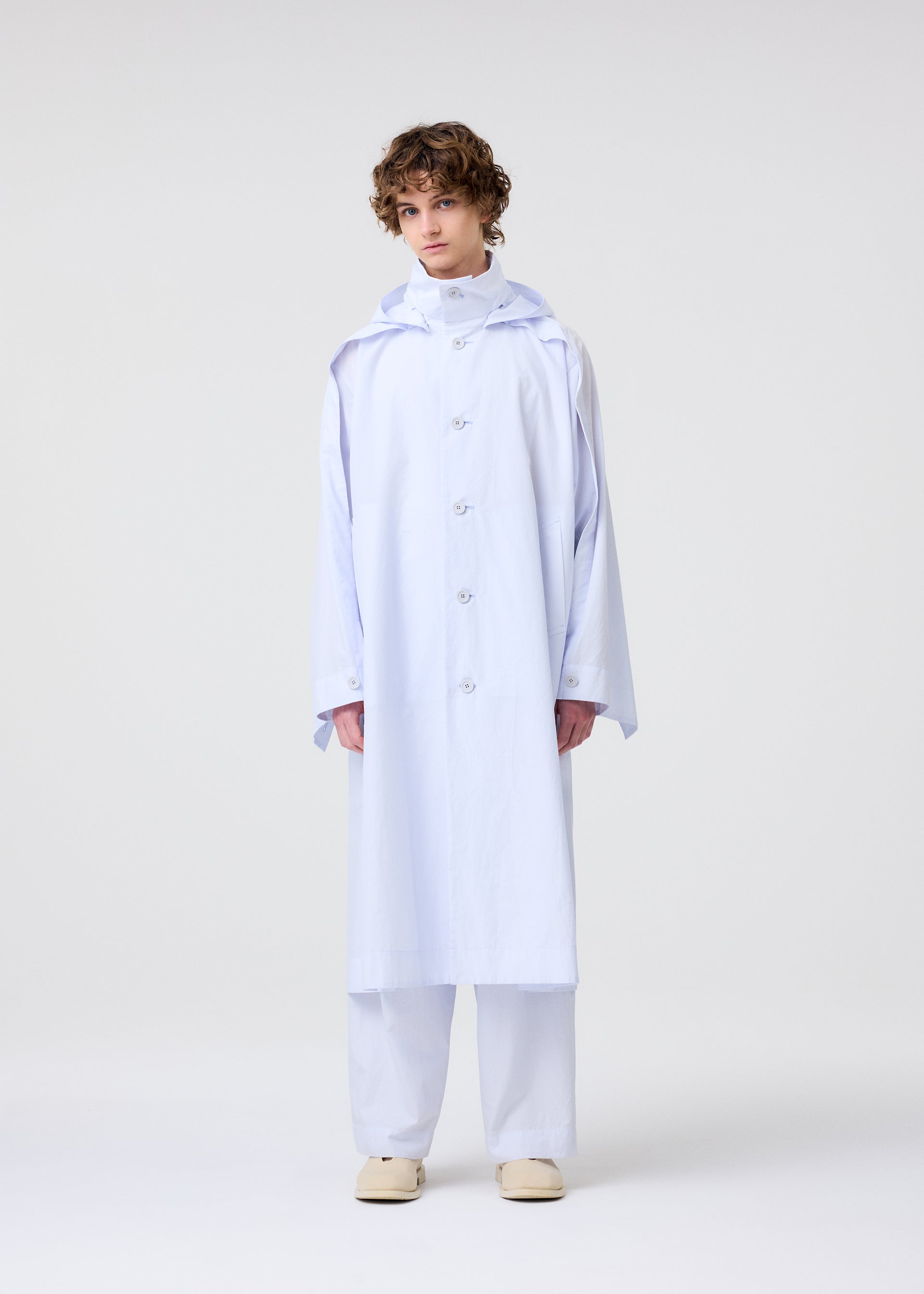 Zoomed view A person with short curly hair wears the FRESHEN hooded coat and matching wide-legged pants by HOMME PLISSÉ ISSEY MIYAKE, styled with beige shoes against a plain background.