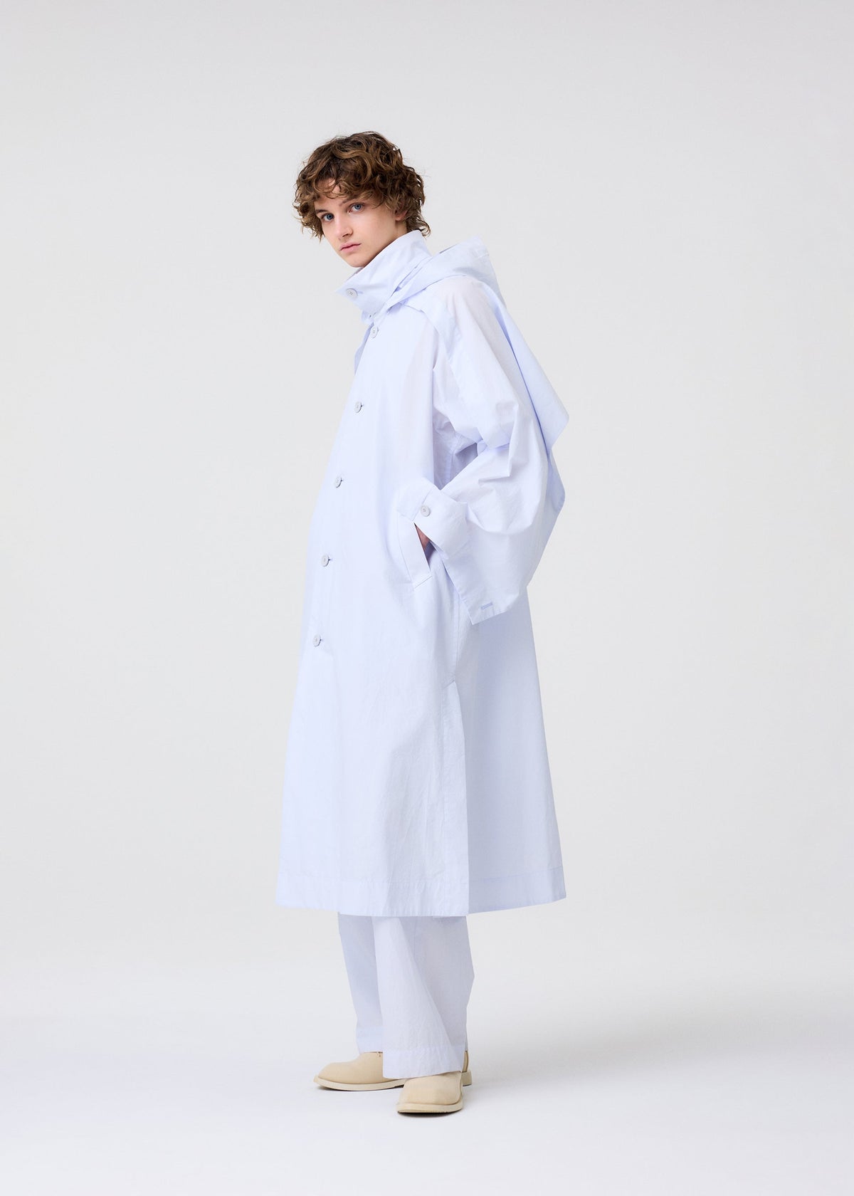 A person with curly hair models the FRESHEN set by HOMME PLISSÉ ISSEY MIYAKE: a loose, light blue wrinkled trench coat with large sleeves and matching pants. With hands in pockets and beige shoes, they stand before a plain light background. view 8 of 9
