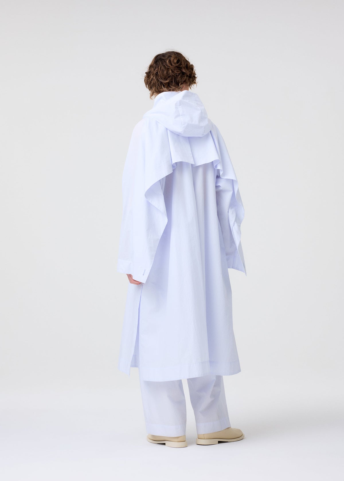 A person with short curly hair faces away, wearing the HOMME PLISSÉ ISSEY MIYAKE FRESHEN light blue hooded coat and matching wide-leg pants with a subtle wrinkled texture, paired with cream shoes against a plain white background. view 9 of 9