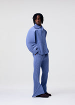 A person with shoulder-length hair and a serious expression models the HOMME PLISSÉ ISSEY MIYAKE LINEN LIKE set: a blue pinstriped outfit with oversized, rounded-sleeve jacket and flared pants, against a plain light background.