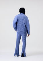 A person with short, curly hair faces away wearing the HOMME PLISSÉ ISSEY MIYAKE LINEN LIKE set—a loose, light blue pinstriped outfit with a linen-like jacket and flared pants with back slits—against a plain light background.